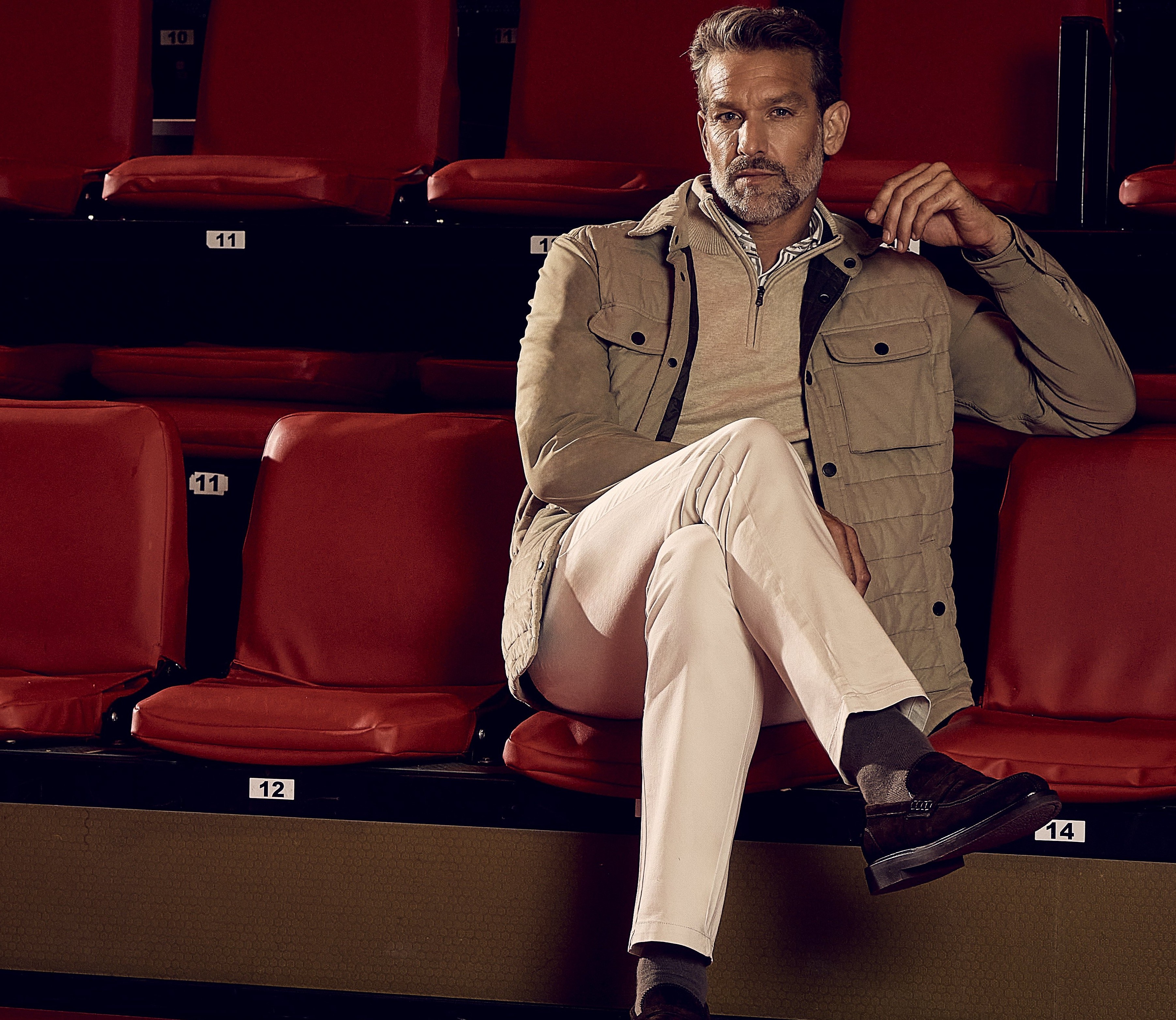 Man seated on red theater seats, wearing a beige jacket, light-colored shirt, and white pants, legs crossed, looking to the side.