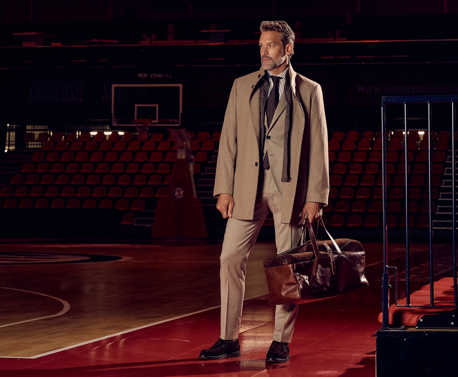 A man in a beige coat and suit stands on a basketball court, holding a leather duffel bag. The stadium seats are empty in the background.