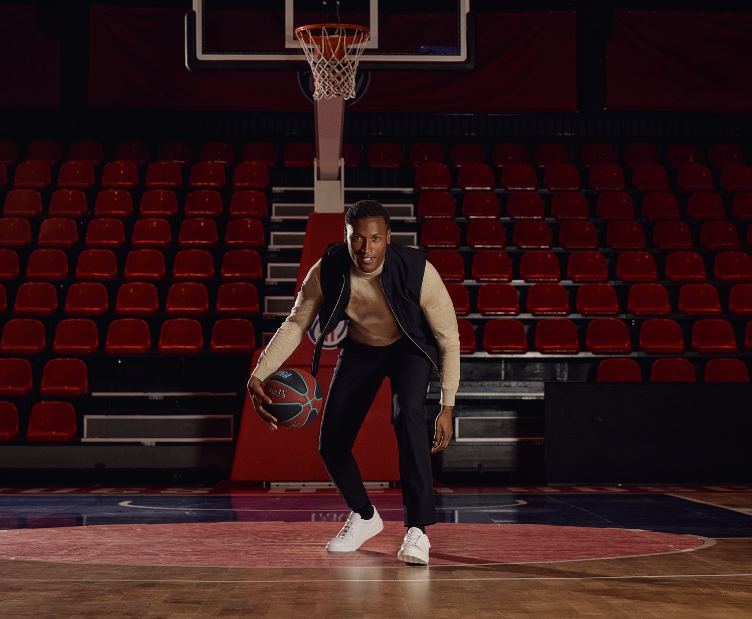 A person dribbles a basketball on an indoor court with empty red seats in the background.