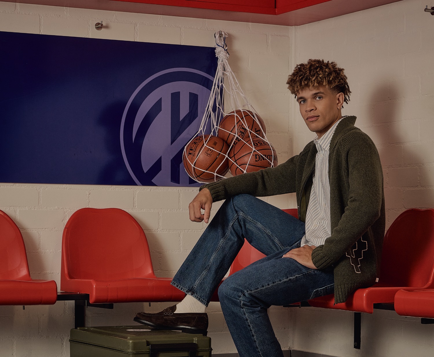 A person sitting on red chairs in a locker room, with basketballs in a net hanging on the wall and a blue sign in the background.