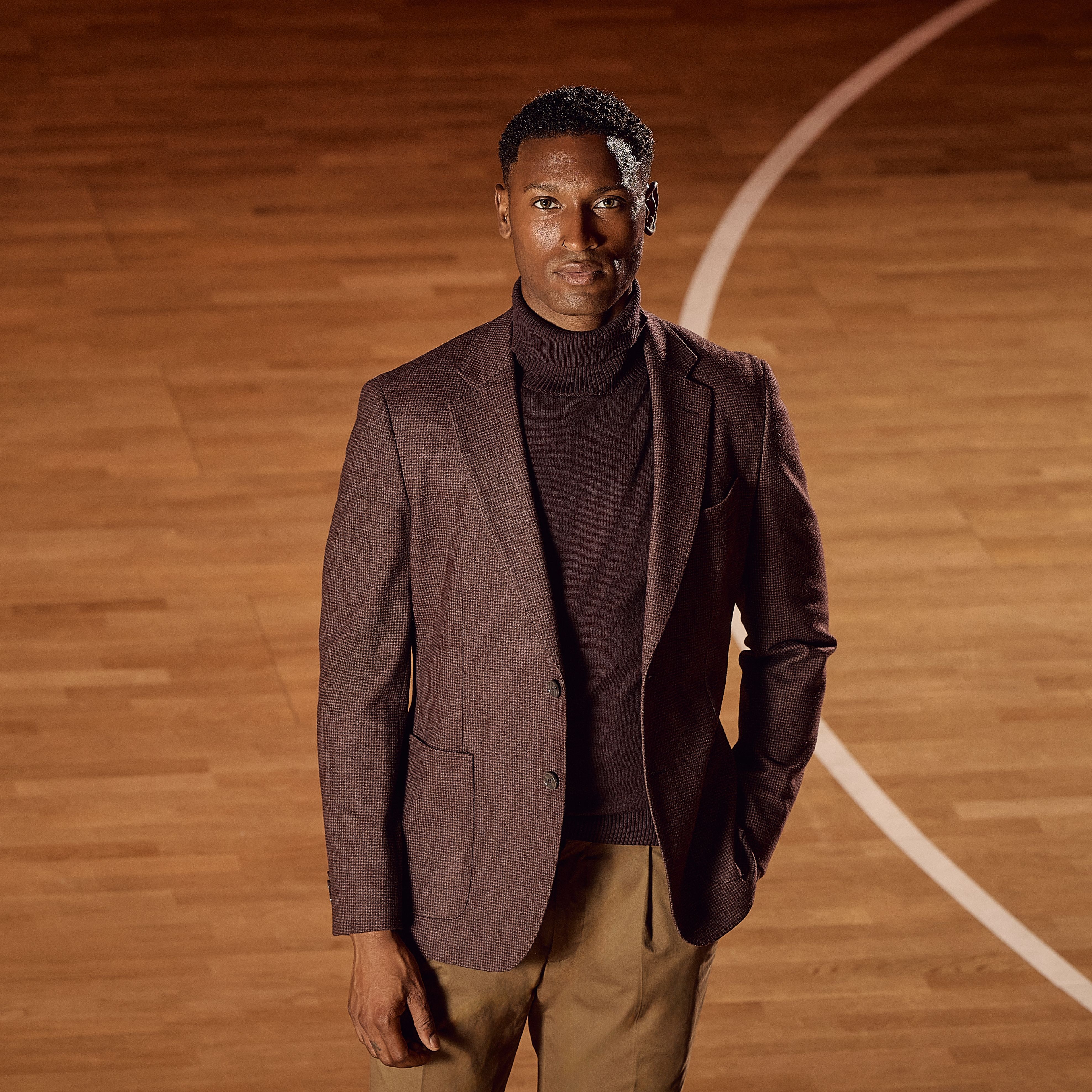 Man in a maroon blazer and brown turtleneck stands on a wooden basketball court, looking confidently at the camera.