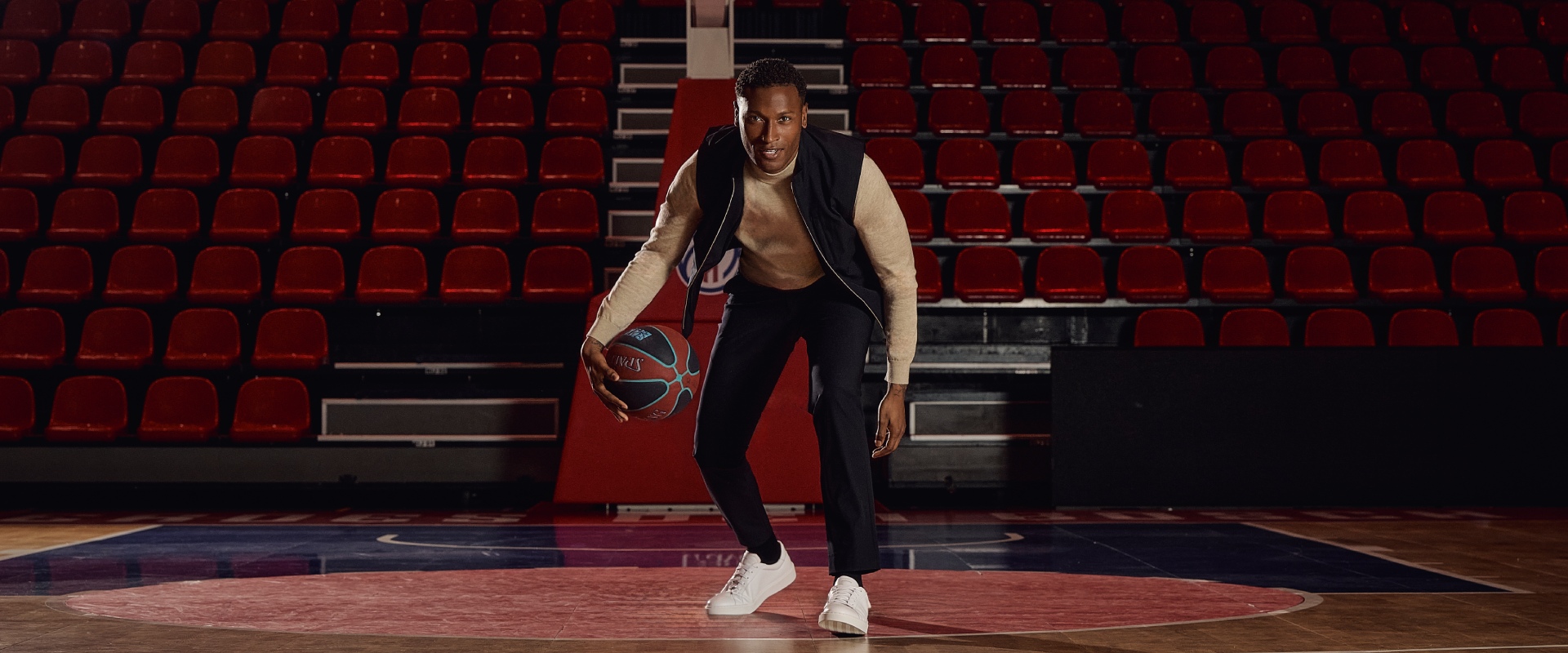 A person dribbles a basketball on an empty indoor court with red seating in the background.