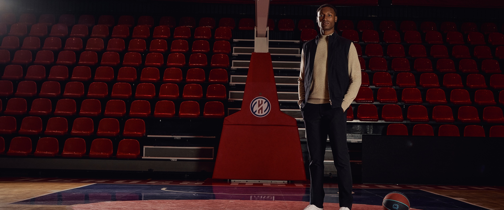 A person stands on a basketball court in front of empty red seats and a basketball hoop, with a basketball on the floor nearby.