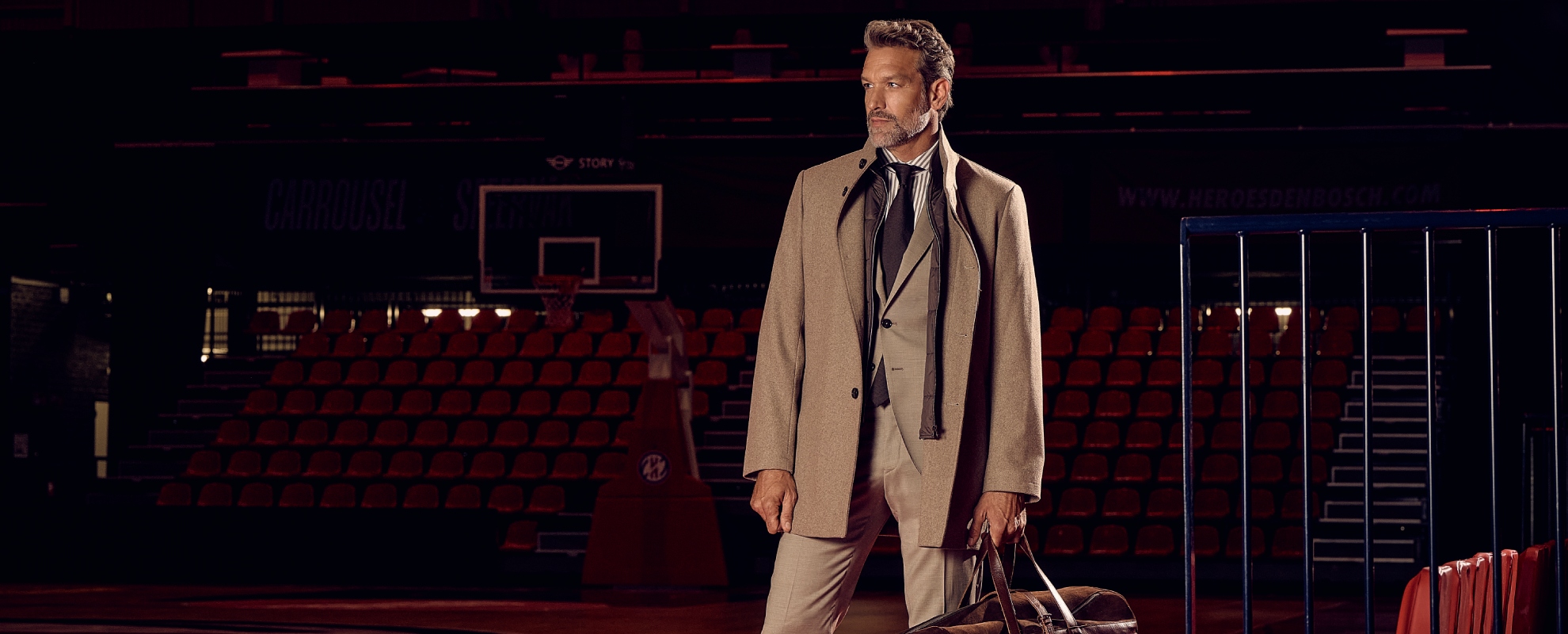A man in a beige suit stands in a dimly lit basketball court holding a bag, with rows of red seats and a basketball hoop in the background.