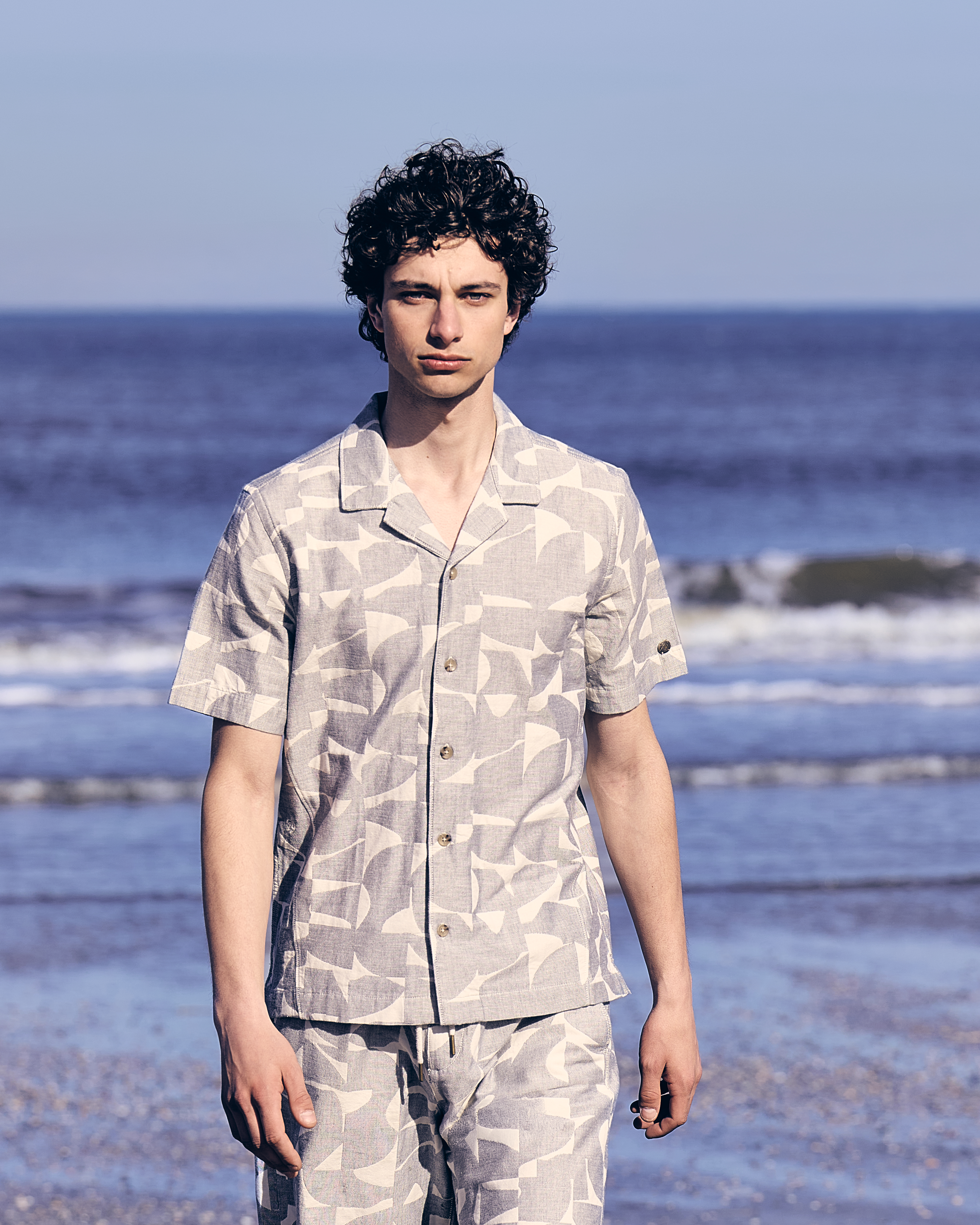 Man in een co-ord op een strand met water op de achtergrond.