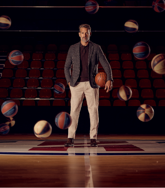 A man in a blazer holds a basketball on a court, surrounded by multiple basketballs in motion, with empty red seats in the background.