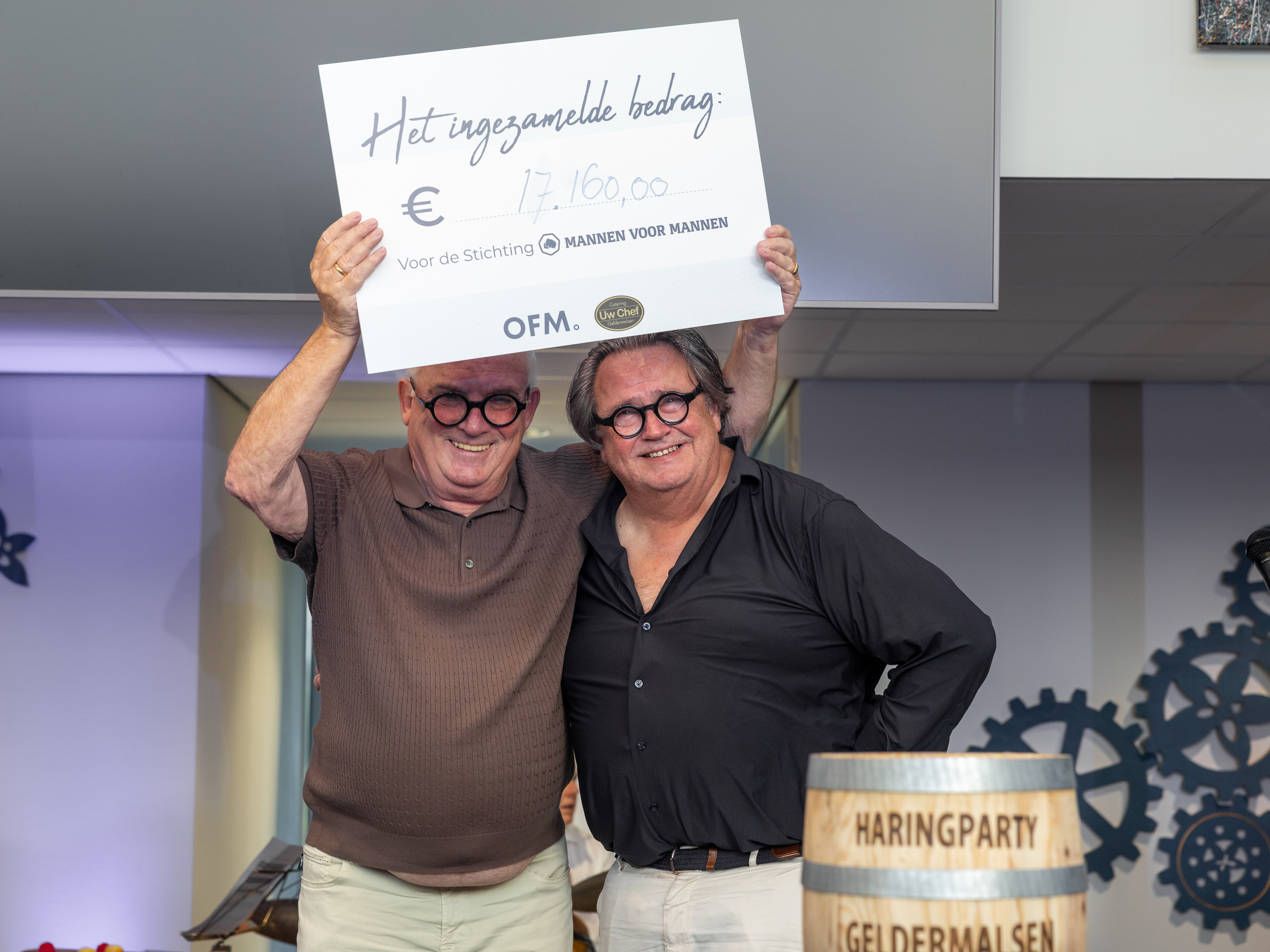 Two smiling men celebrate, holding a large check for €17,160.00 at an event with a "Haringparty" barrel in the foreground.
