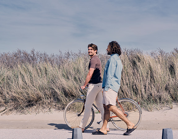 Two people walking on a beach path; one pushing a bike. Tall grasses and a blue sky form the background.