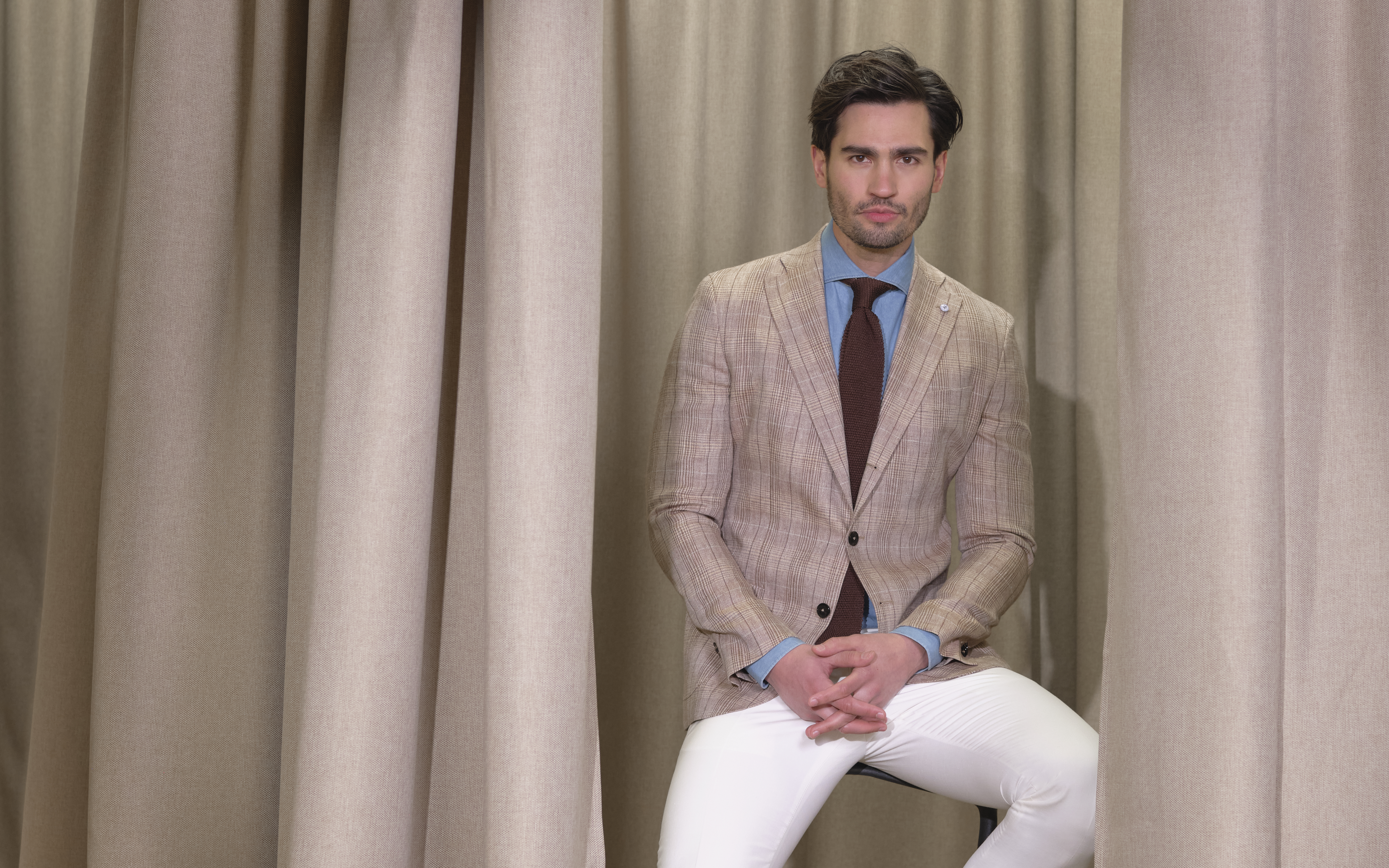 Man in a beige blazer, blue shirt, and white pants sits on a stool against a backdrop of beige curtains.