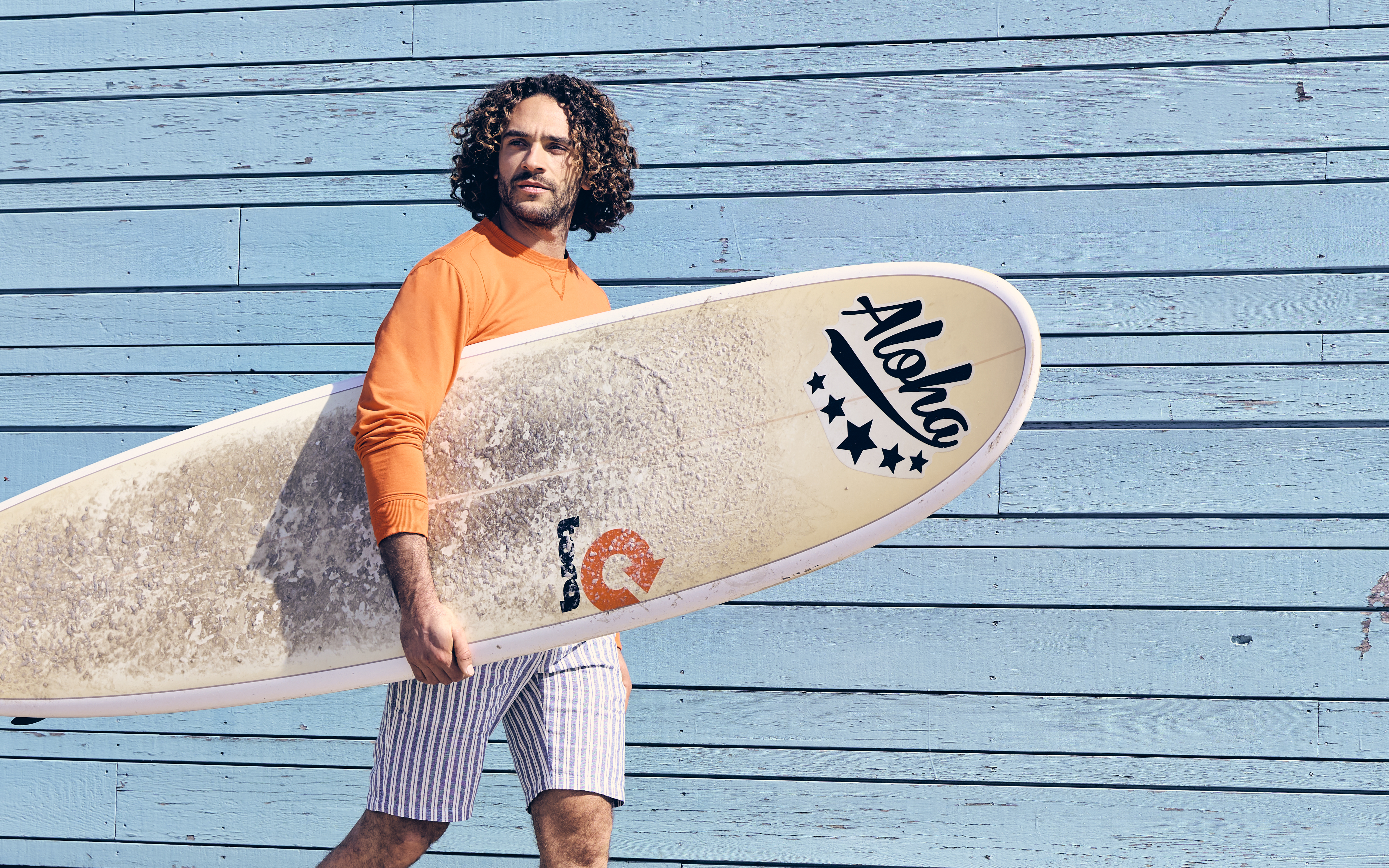 Man in an orange shirt holding a surfboard with "Aloha" text, standing in front of a blue wooden wall.