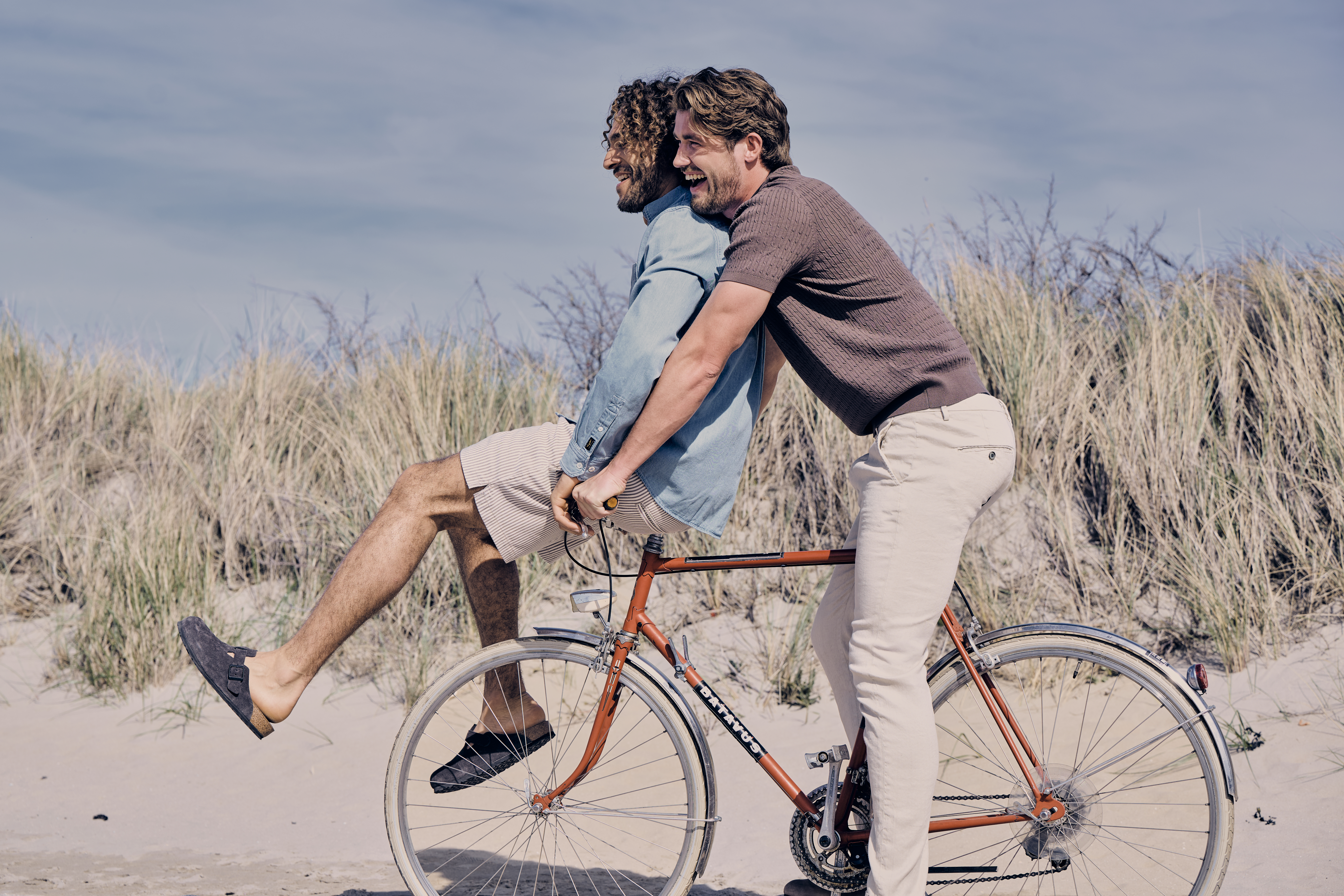 Two people enjoy a bike ride on a sandy path near tall grass, with one sitting on the handlebars and the other pedaling. They are wearing summeroutftits.