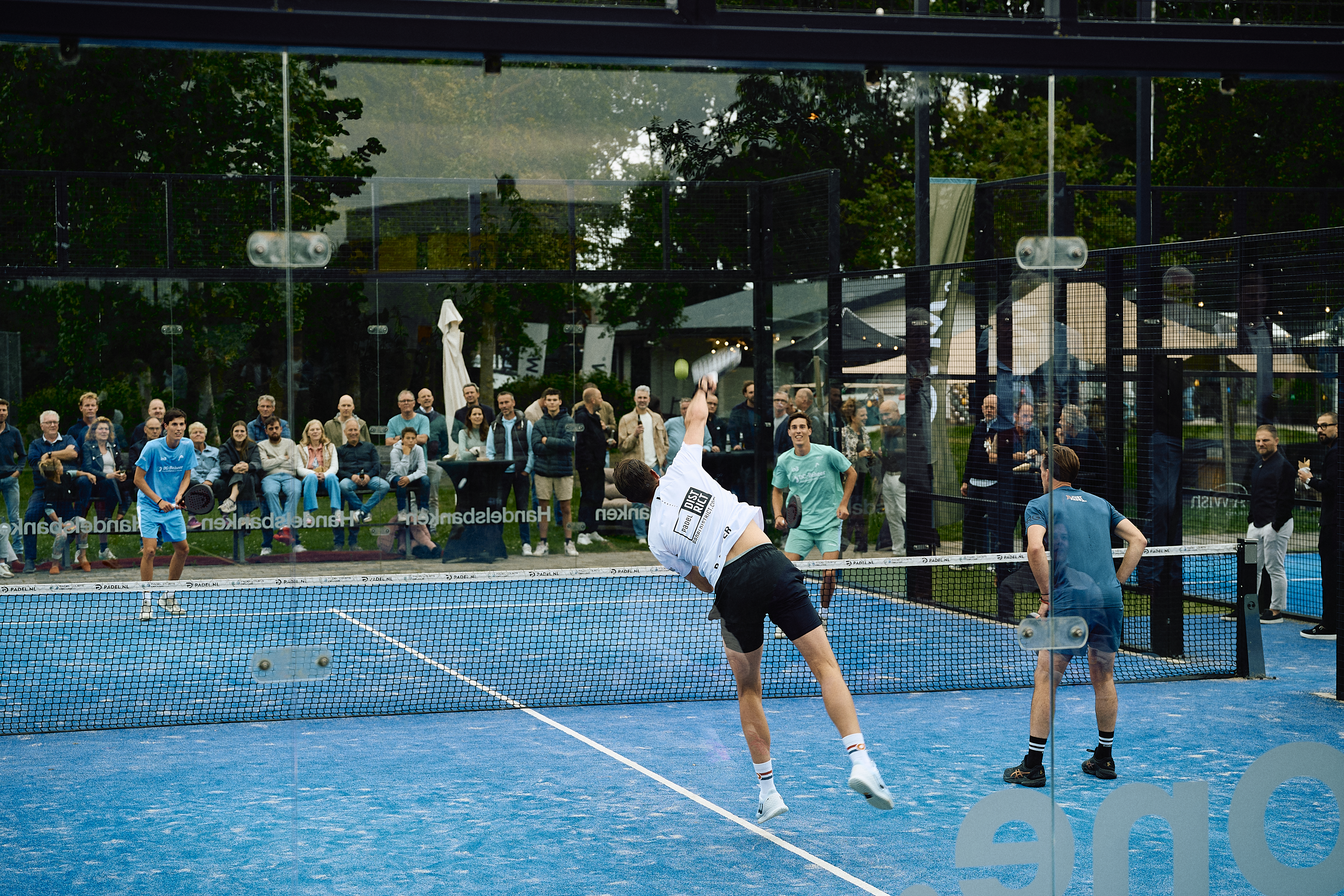 Two players compete in a doubles padel match on a blue court, with a cheering crowd in the background.