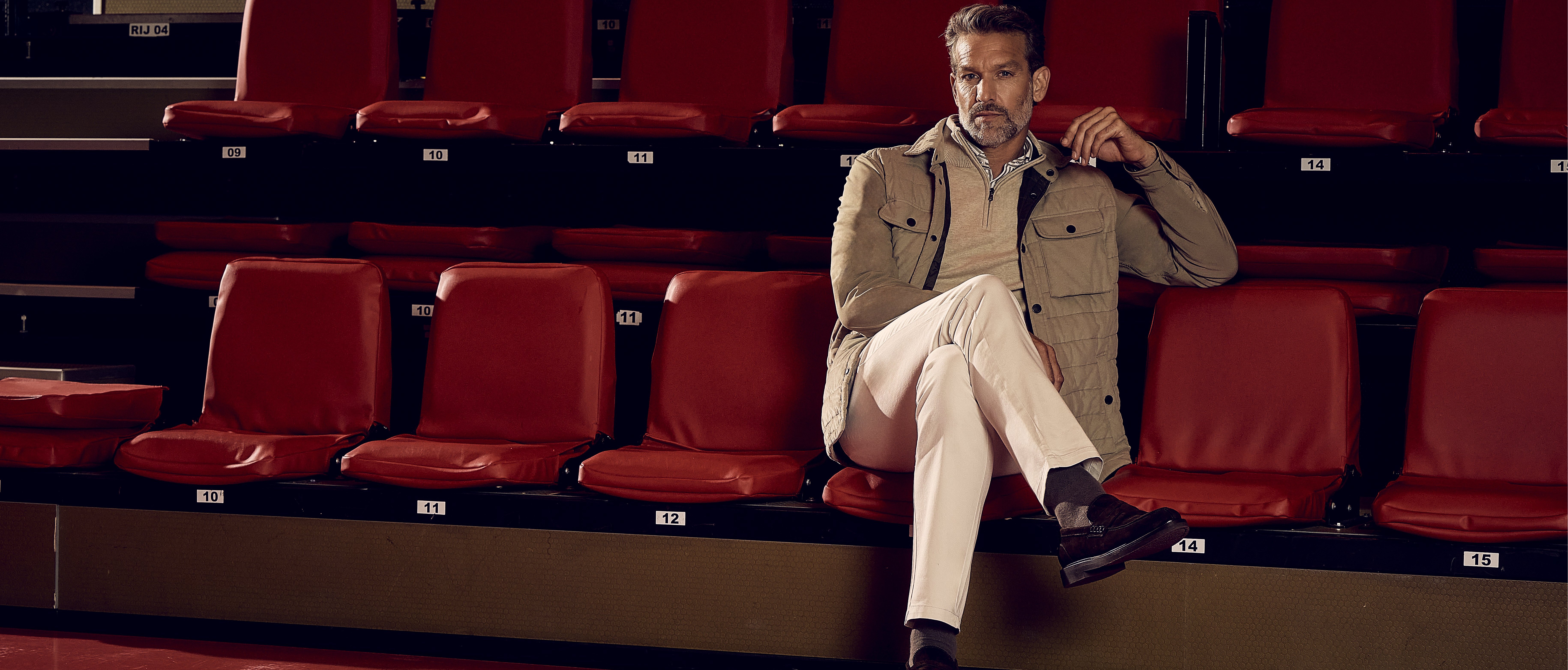 Man in a beige jacket and white pants sits on red auditorium seats, with legs crossed and thoughtful expression.