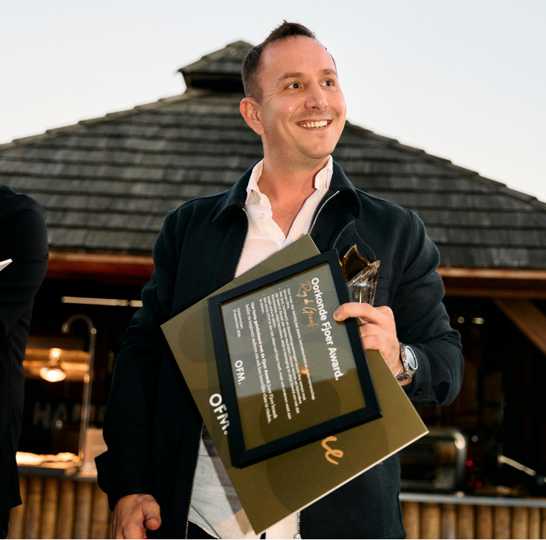 Man smiling outdoors, holding an "Outstanding Paper Award" certificate and a trophy, wearing a dark jacket against a wooden building background.