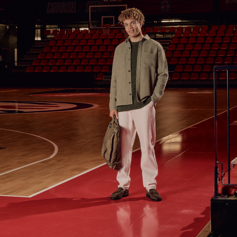 Smiling person standing on a basketball court, wearing a green jacket and white pants, holding a bag with their hands in pockets.