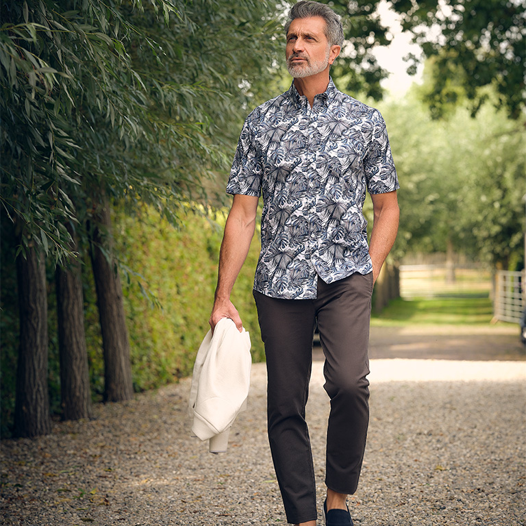 Man gekleed in een bruine chino en blauw gebloemd overhemd wandelend over een pad met een wit vest in zijn hand