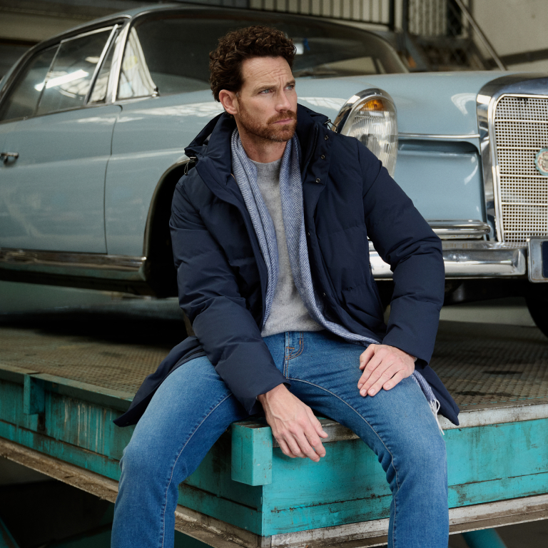 Man gekleed in donkerblauwe jas, grijze trui en blauwe jeans.