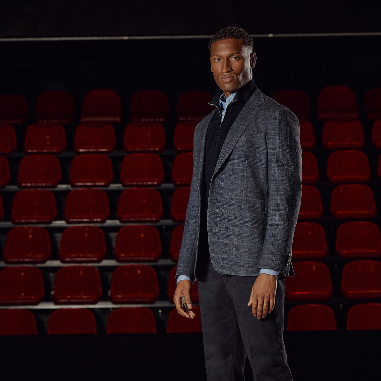 A person in a gray blazer stands confidently in front of empty red auditorium seats, with a serious expression and direct gaze.