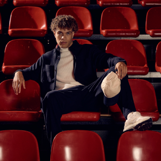 A person with curly hair, dressed in a dark jacket, light sweater, and pants with a light patch, sits confidently on red stadium seats.