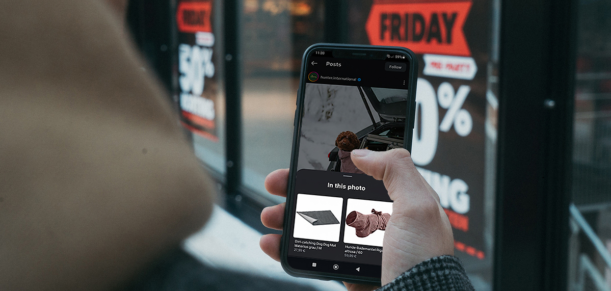 Person holding a smartphone displaying an online shopping app with Black Friday sale signs, offering 50% off, visible in the background.