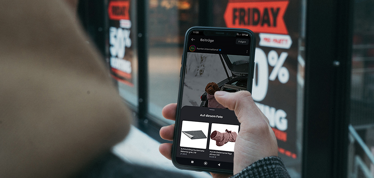 Eine Person hält ein Smartphone in der Hand, auf dem eine Online-Shopping-App mit Black-Friday-Angeboten zu sehen ist; im Hintergrund sind Schilder mit ‚50 % Rabatt‘ erkennbar.