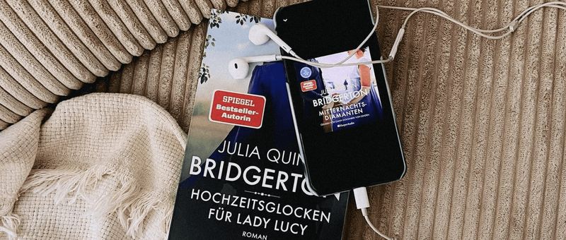 Ein Buch mit dem Titel „Bridgerton“ von Julia Quinn liegt auf einer Couch neben einem Smartphone mit Kopfhörern, was auf ein Hörbuch oder Musik schließen lässt.