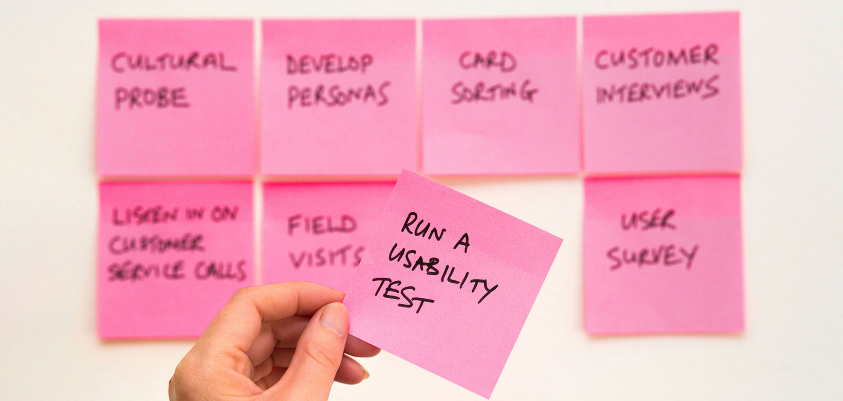 Eine Hand, die einen rosa Haftzettel mit der Aufschrift „Führe einen Usability-Test durch“ hält, im Hintergrund sind weitere Zettel an einer Wand zu sehen, auf denen verschiedene Forschungsmethoden aufgeführt sind.