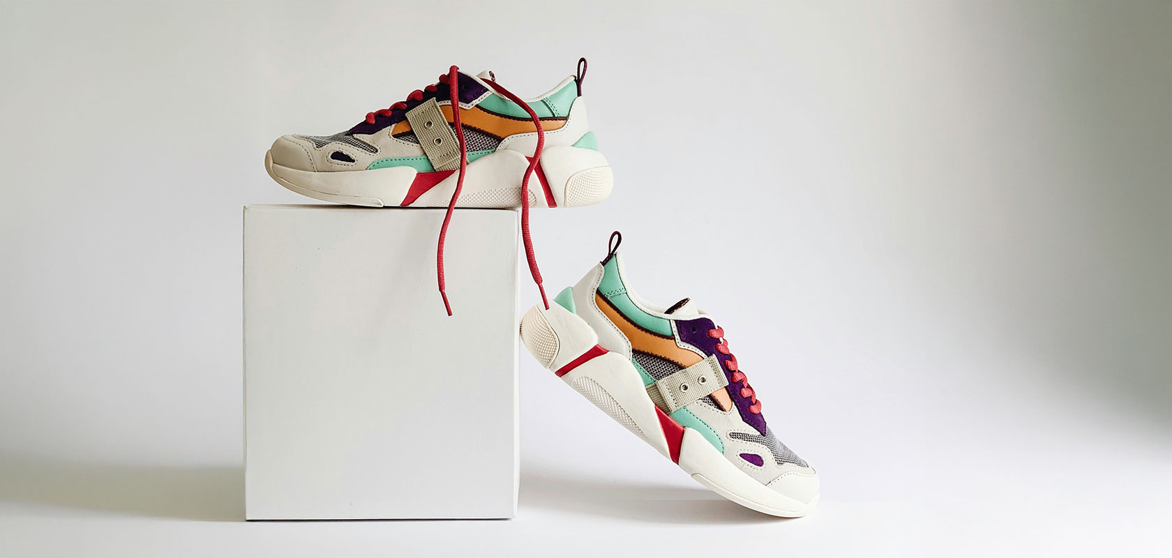 Ein Paar mehrfarbige Sneaker, präsentiert auf und neben einem weißen Würfel vor hellem Hintergrund.