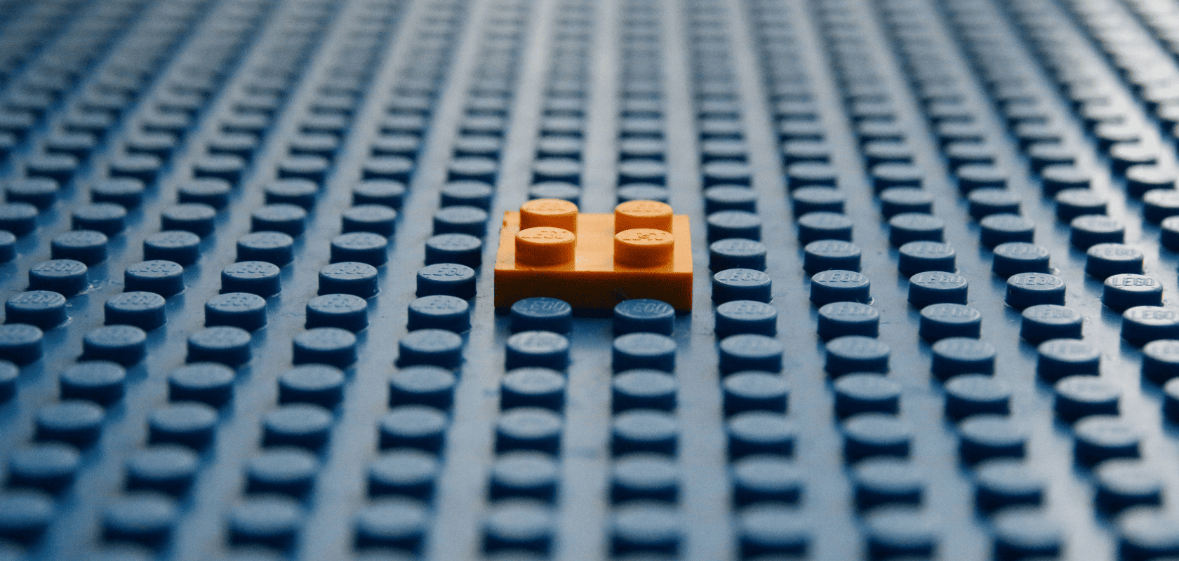 A small orange LEGO brick on a large blue baseplate with a grid of round studs, viewed from a low angle.