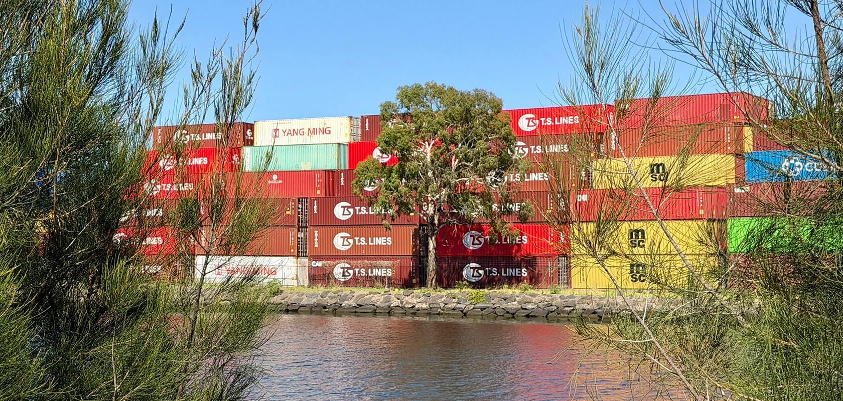 An einer Uferpromenade gestapelte Schiffscontainer, eingerahmt von Bäumen, mit strahlend blauem Himmel im Hintergrund.