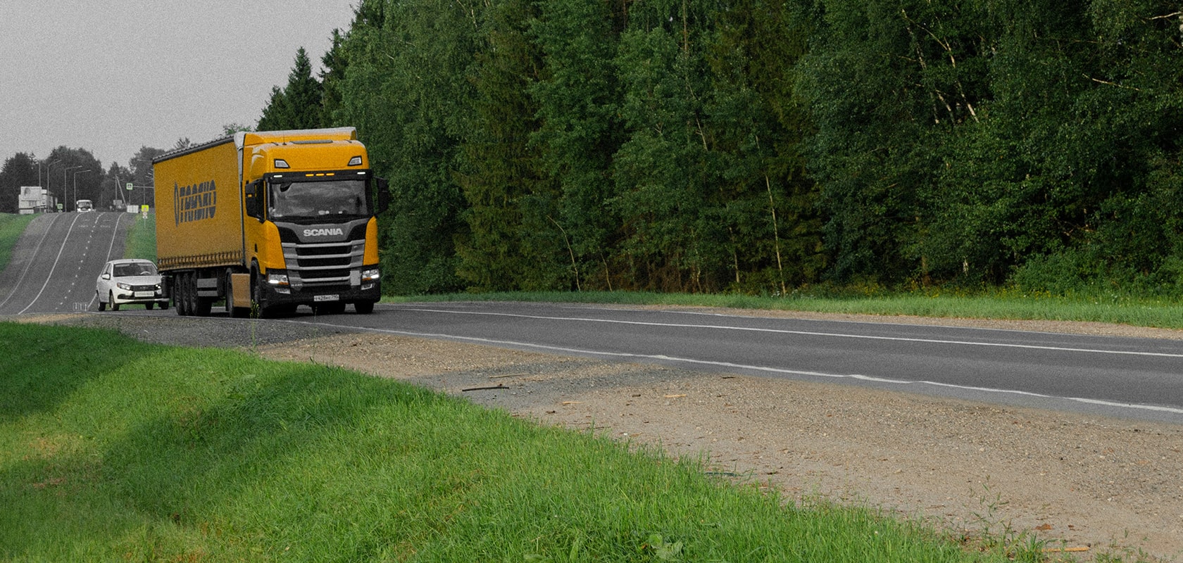 Gelber Lastwagen und weißes Auto fahren auf einer Landstraße, die von grünen Bäumen und Gras gesäumt ist, unter einem bewölkten Himmel.