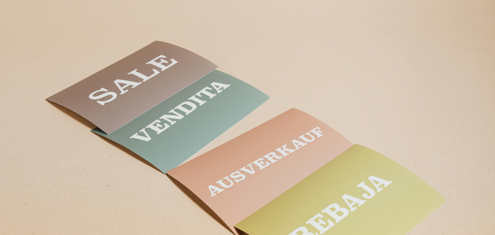 Schilder mit der Aufschrift „Sale“ auf Englisch, Italienisch, Deutsch und Spanisch auf farbigem Papier, angeordnet auf einem beigen Untergrund.