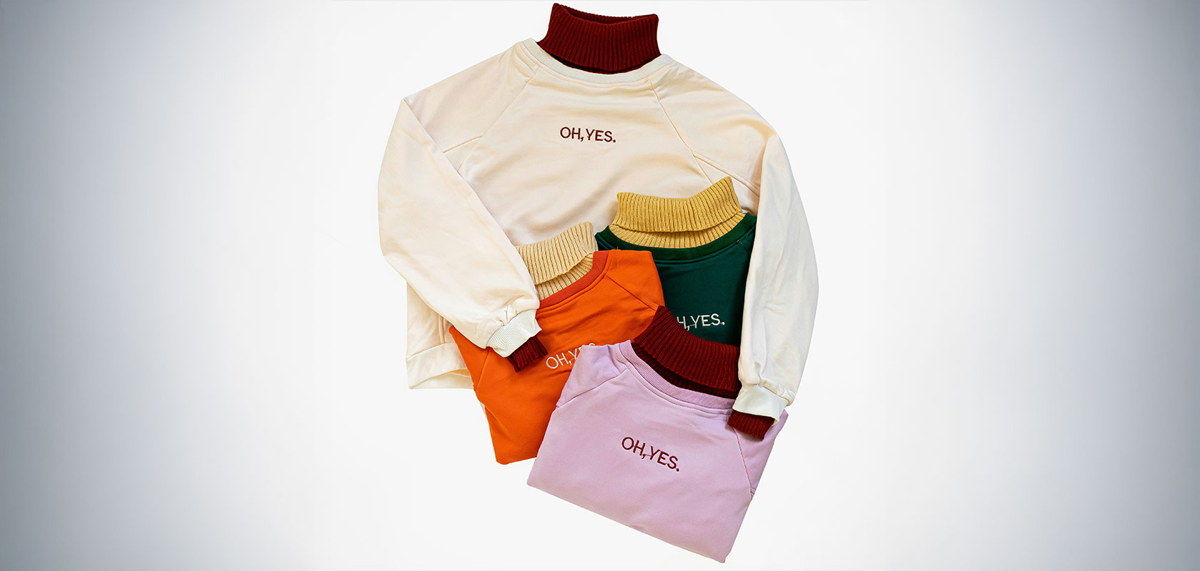 Assorted sweatshirts in cream, orange, green, and pink, each with "OH YES." printed, displayed overlapping on a white background.