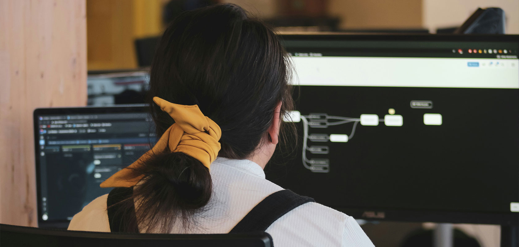Person mit langen, mit einem gelben Band zusammengebundenen Haaren, die an zwei Computermonitoren mit Flussdiagrammen in einer Büroumgebung arbeitet.