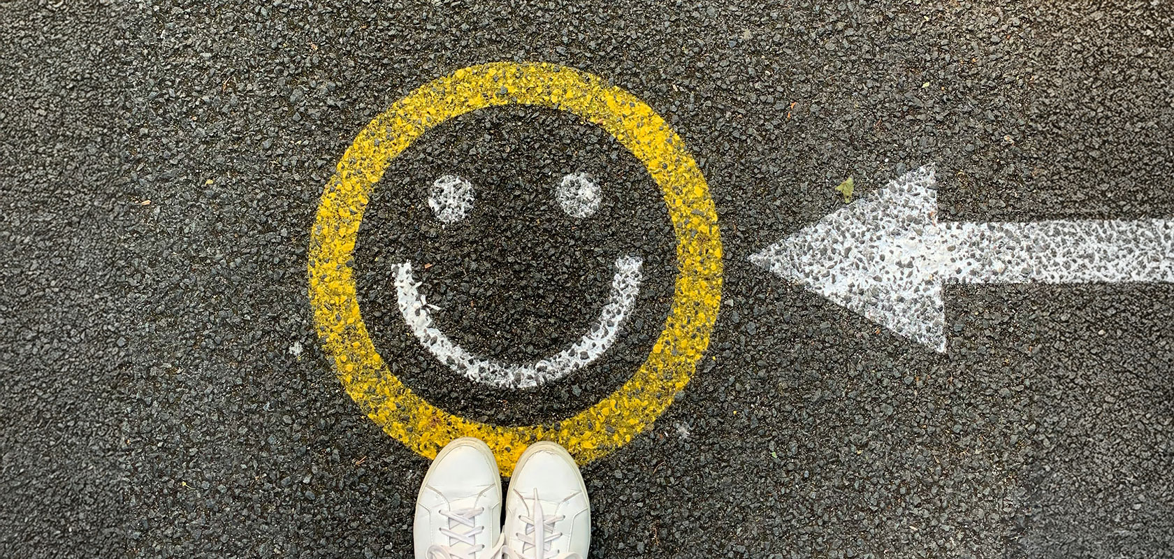 Weiße Turnschuhe auf Asphalt neben einem gelb gemalten Smiley in einem Kreis, mit einem weißen Pfeil, der nach links zeigt.