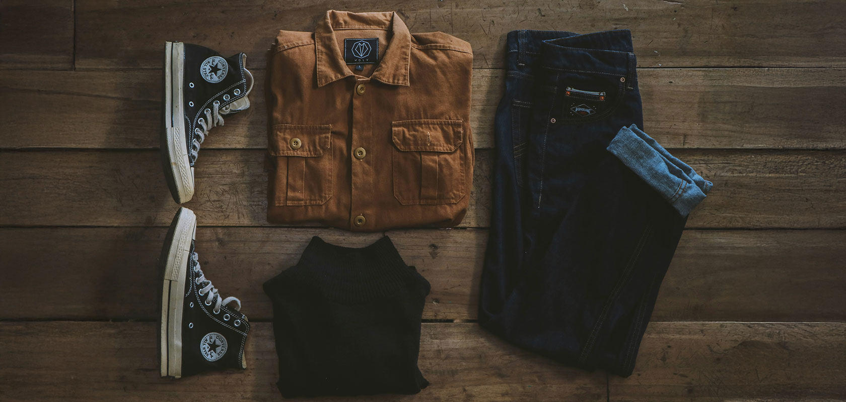 Casual outfit on wooden floor: brown jacket, black shoes with white soles, black sweater, and blue jeans with cuffs rolled.