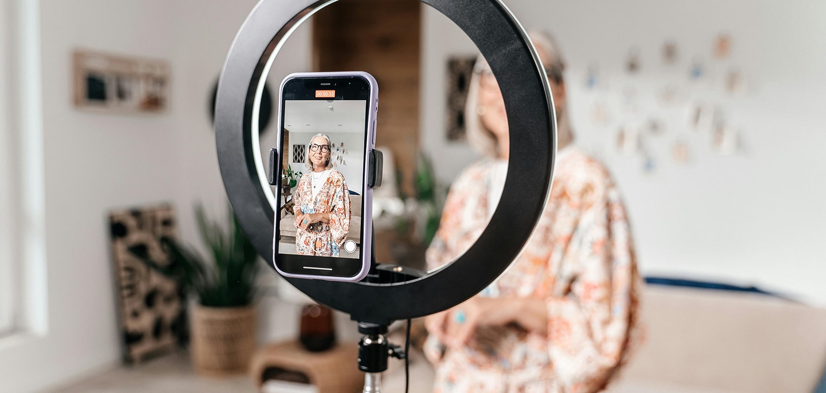 Eine Person, die mit einem Smartphone auf einem Ringlichtstativ in einem hell beleuchteten Raum gefilmt wird, wobei im Hintergrund verschwommene Einrichtungsgegenstände zu sehen sind.
