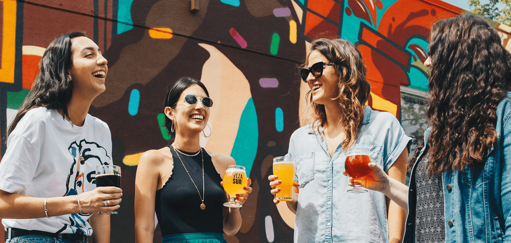 Four people laughing and holding drinks in front of a colorful mural, enjoying a sunny day outdoors.