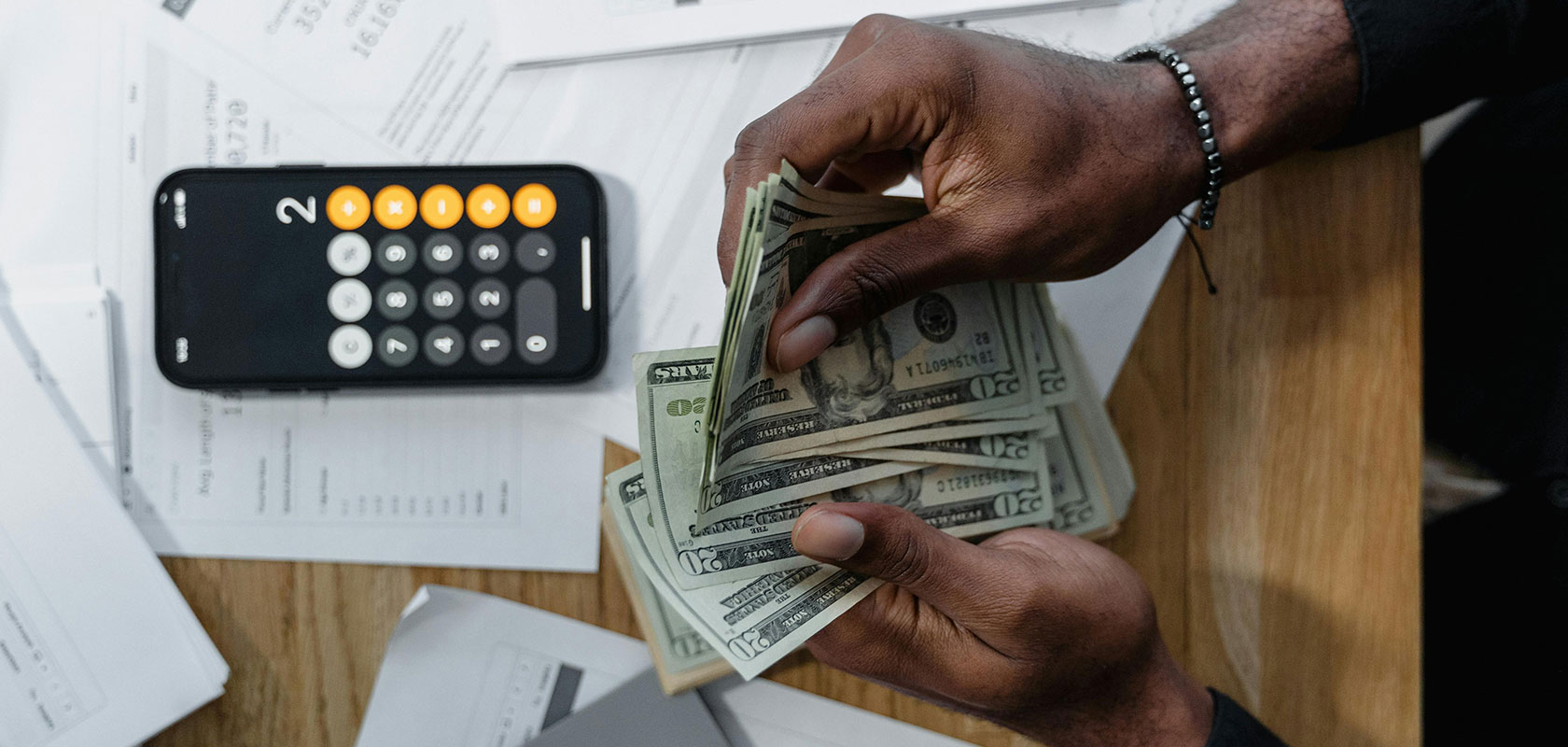 Hände, die US-Dollar auf Dokumenten zählen, neben einem Smartphone mit Taschenrechner.