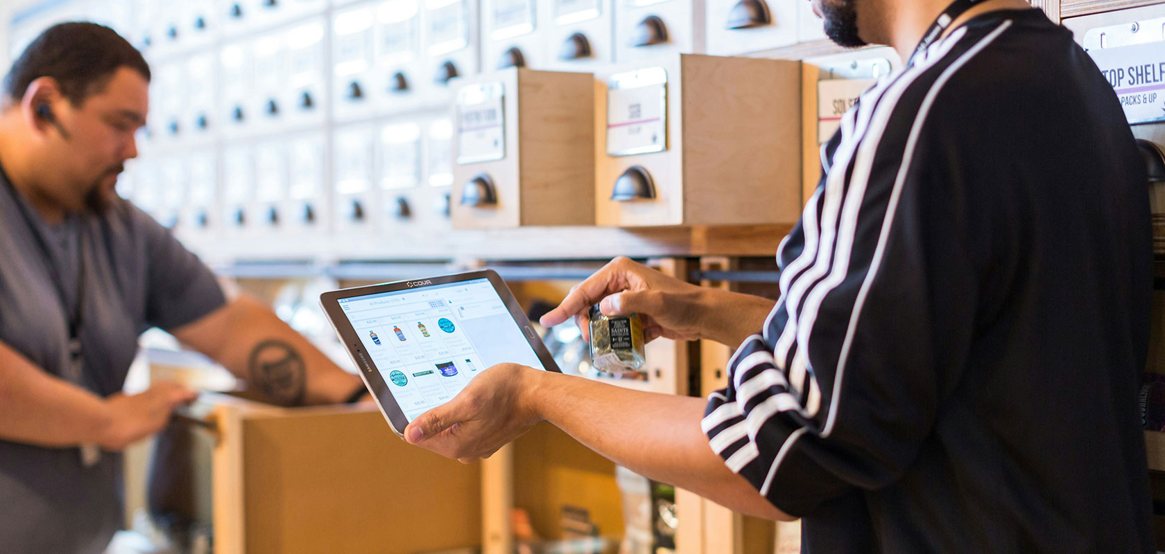 Two people organizing inventory in a storage area; one uses a tablet while the other checks items, with labeled drawers in the background.