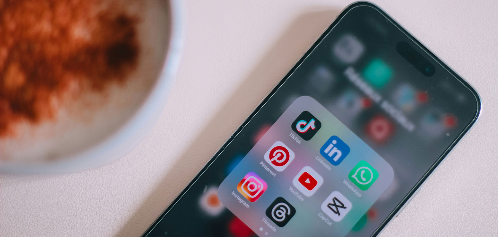 Smartphone mit Symbolen für Social-Media-Apps wie TikTok, Pinterest, Instagram, YouTube, LinkedIn, WhatsApp und anderen neben einer Kaffeetasse.