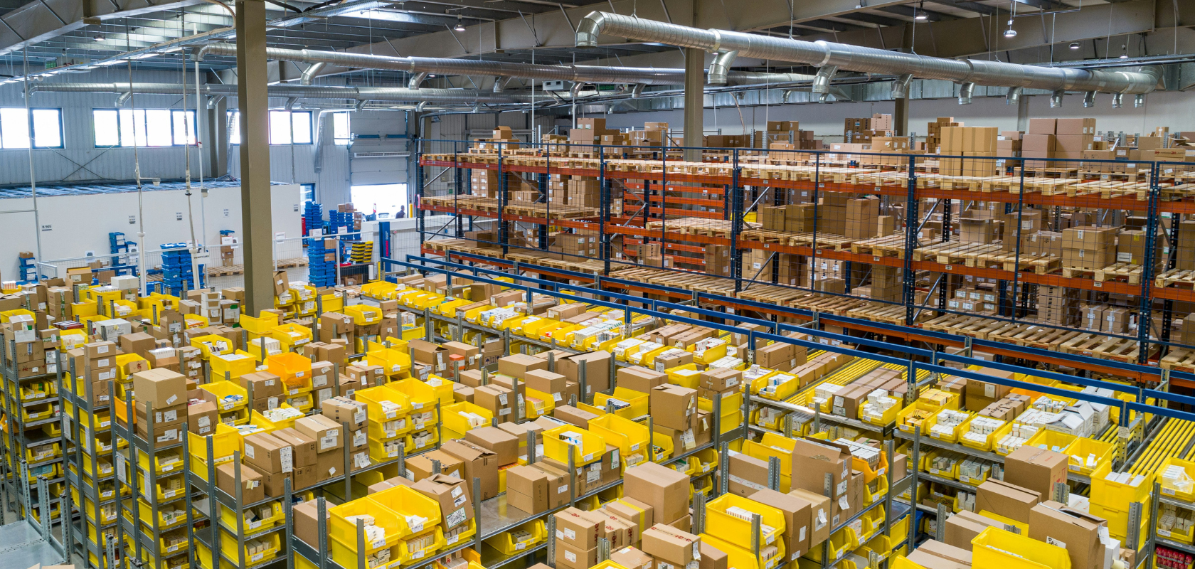 Lagerhalle mit aufgeräumten Regalen, die mit Kisten und Paketen gefüllt sind, unter heller Beleuchtung. Mehrere Reihen gelber und brauner Behälter sind zu sehen.