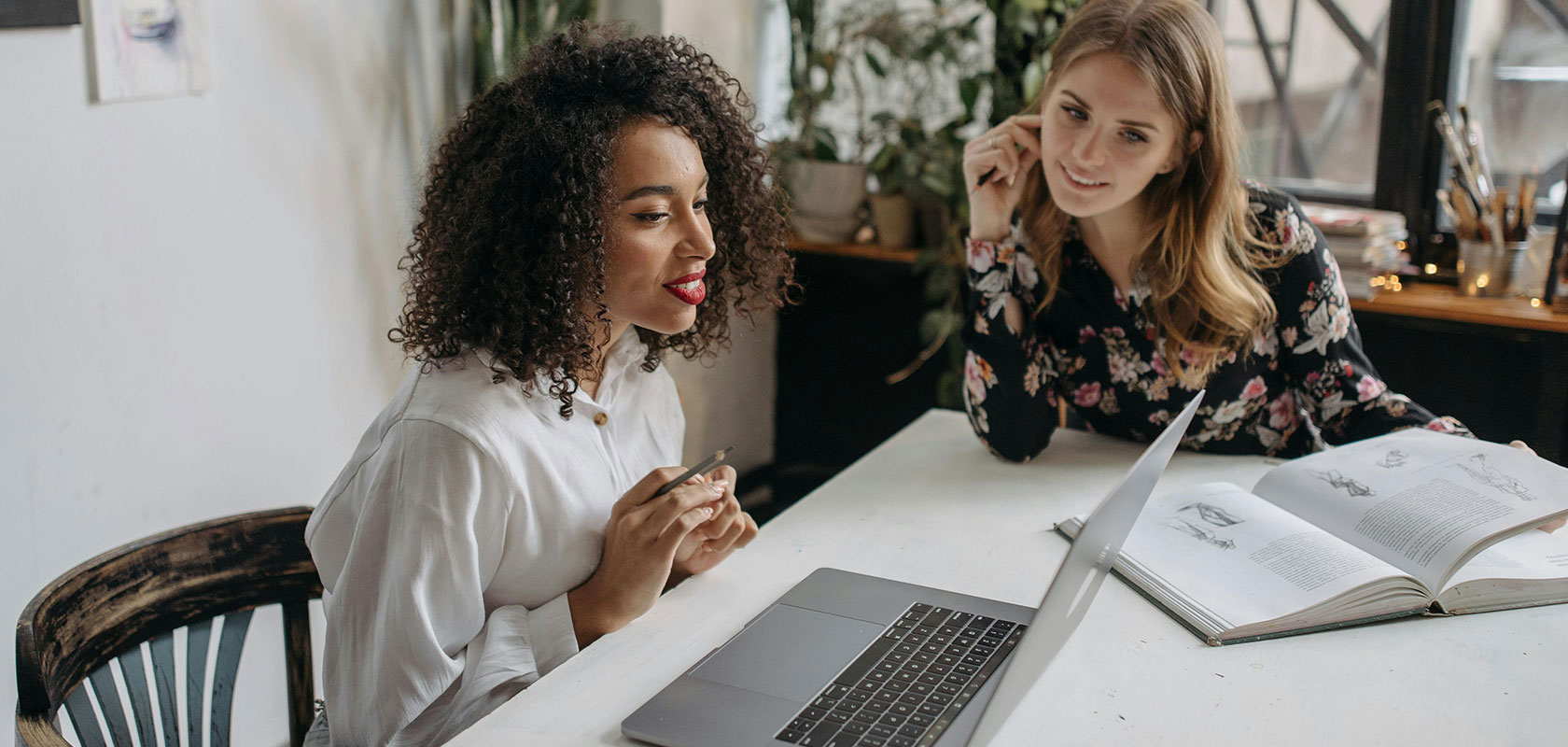 Zwei Frauen sitzen an einem hellen Tisch und arbeiten zusammen; eine spricht, während die andere zuhört, zwischen ihnen liegen ein Laptop und ein aufgeschlagenes Buch.