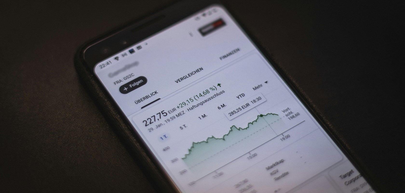 Geneigtes Smartphone auf einer dunklen Oberfläche, das einen Börsenchart mit einem steigenden Liniendiagramm und numerischen Finanzdaten anzeigt.