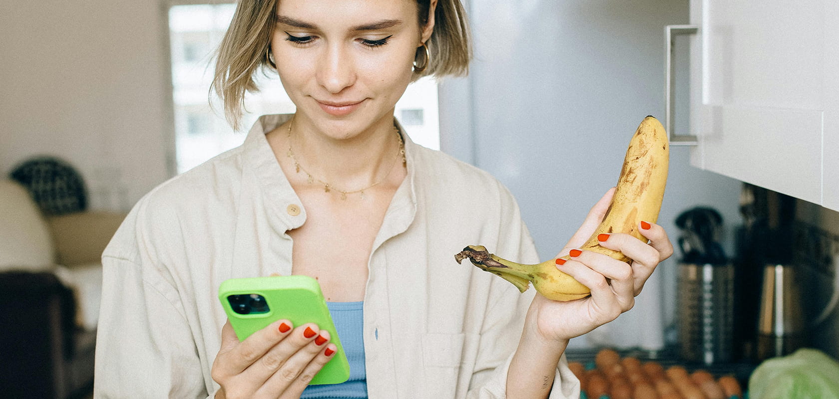 Frau in einer Küche, die eine Banane in der Hand hält und mit konzentriertem Blick auf ihr Handy schaut.