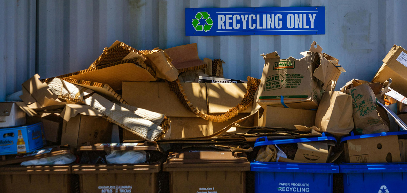 Stapel von Kartons und Papierboxen, die in blauen Recyclingbehältern unter einem Schild mit der Aufschrift „Nur Recycling“ vor einer Wellblechwand gestapelt sind.