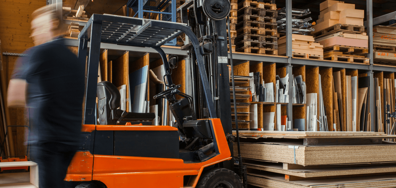 A forklift and an employee moving around in a warehouse with high racks full of wood panels and pallets.