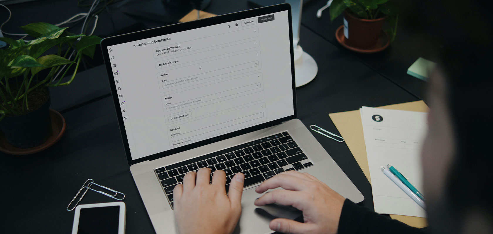 Eine Person, die auf einem Laptop tippt, auf dem ein Dokument geöffnet ist, umgeben von Pflanzen, Papieren und Schreibutensilien auf einem Schreibtisch.