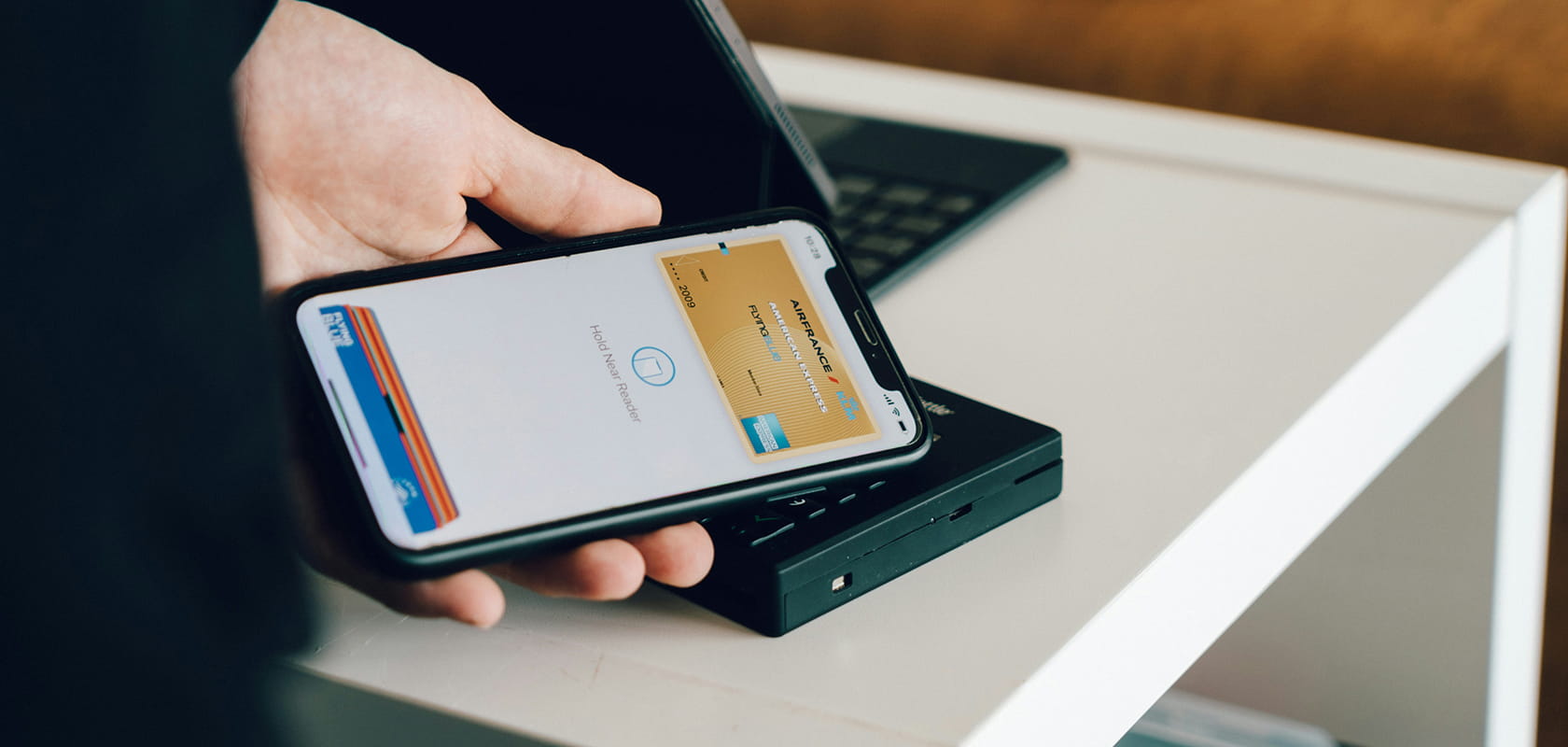 Person, die ein Smartphone in der Nähe eines Zahlungsterminals hält und bereit ist, eine kontaktlose Transaktion durchzuführen. Im Hintergrund ein Laptop auf einem weißen Tisch.