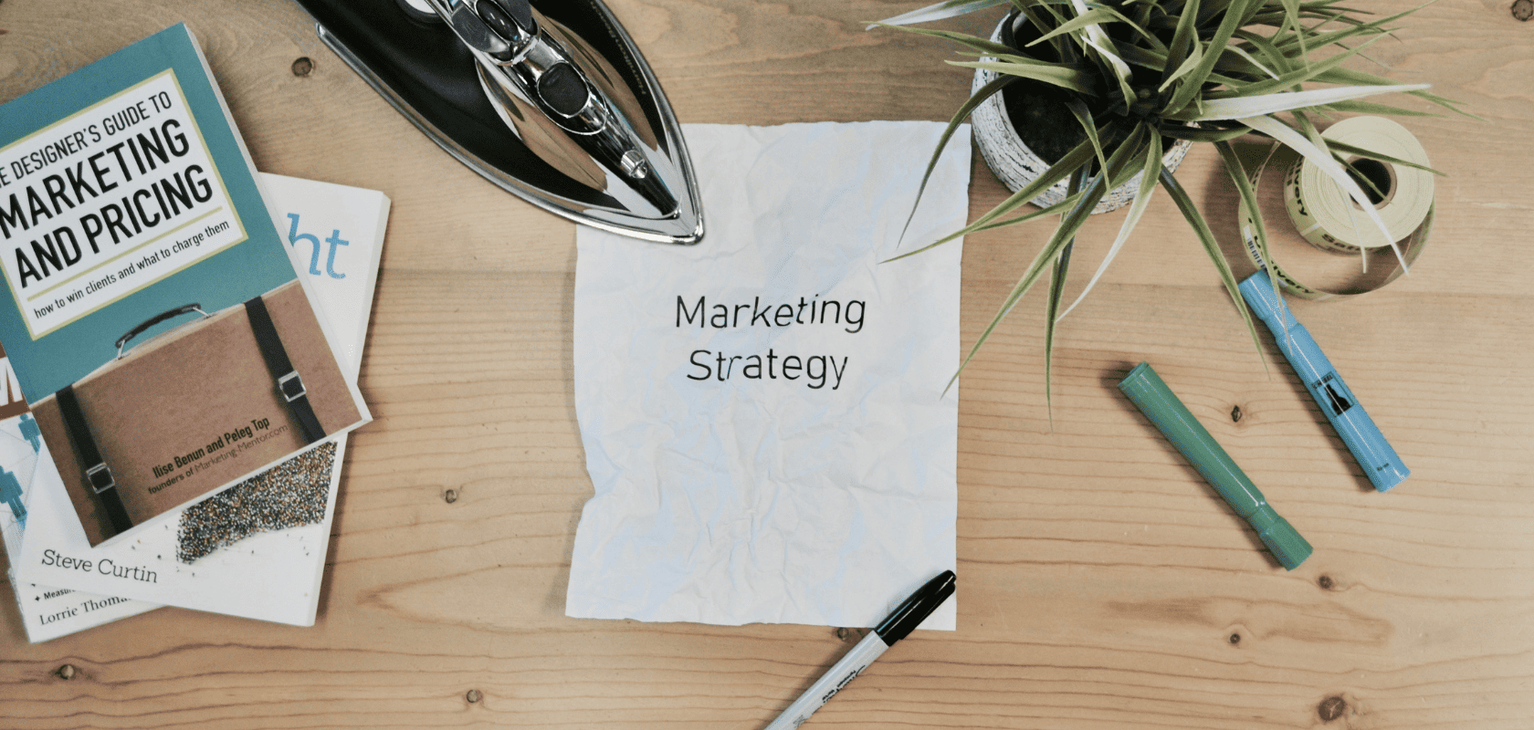 A folded sheet of paper labeled “Marketing Strategy” on a wooden desk, surrounded by an iron, reference books, felt-tip pens, and a potted plant.